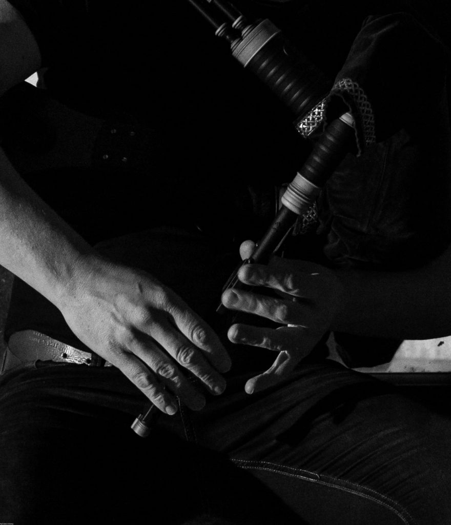 Black and white close-up of a musician playing a Lindsay System bagpipe chanter, focusing on hand positioning and the instrument’s intricate craftsmanship in a moody, artistic composition.