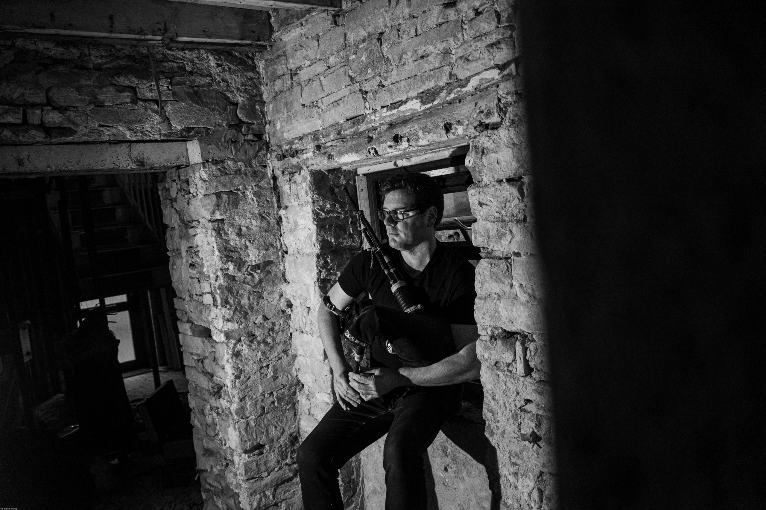 Black and white portrait of Donald Lindsay playing a Lindsay System bagpipe chanter, seated in a stone window frame inside the historic Old Post Office House in St Margaret's Hope, Orkney.