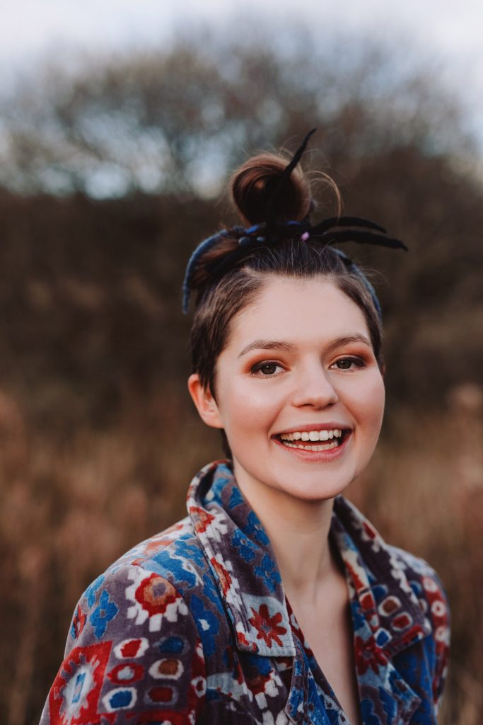 Evie Waddell smiling outdoors in natural light, wearing a colourful patterned jacket with a bold updo tied in yarn and ribbon. A soft-focus background of trees and tall grasses adds an autumnal feel. Photo by Elly Lucas.