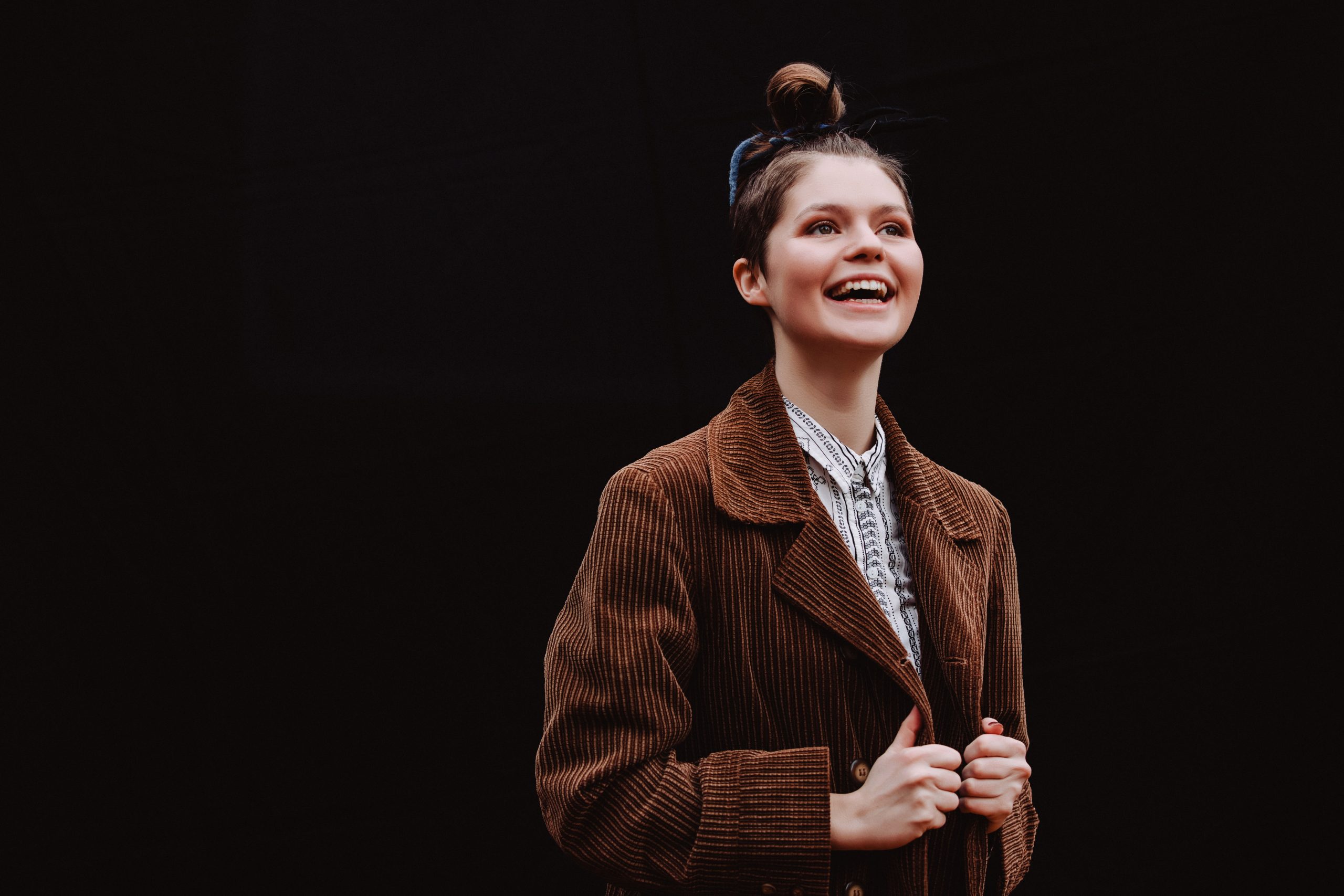 Evie Waddell smiling and looking upwards, wearing a brown corduroy coat and patterned shirt, with her hair tied in a top knot and a ribbon. The background is dark, making her stand out clearly. Photo by Elly Lucas.
