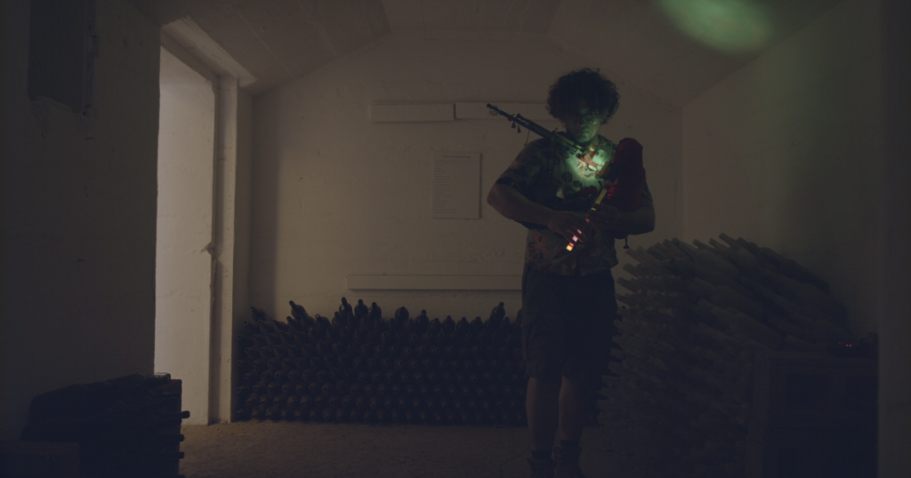 Donald Lindsay performing with his illuminated Lindsay System bagpipe chanter in a bottle room of Fort Hayes, Ascension Island. The dimly lit space highlights the glowing instrument and rows of stacked glass bottles lining the walls.