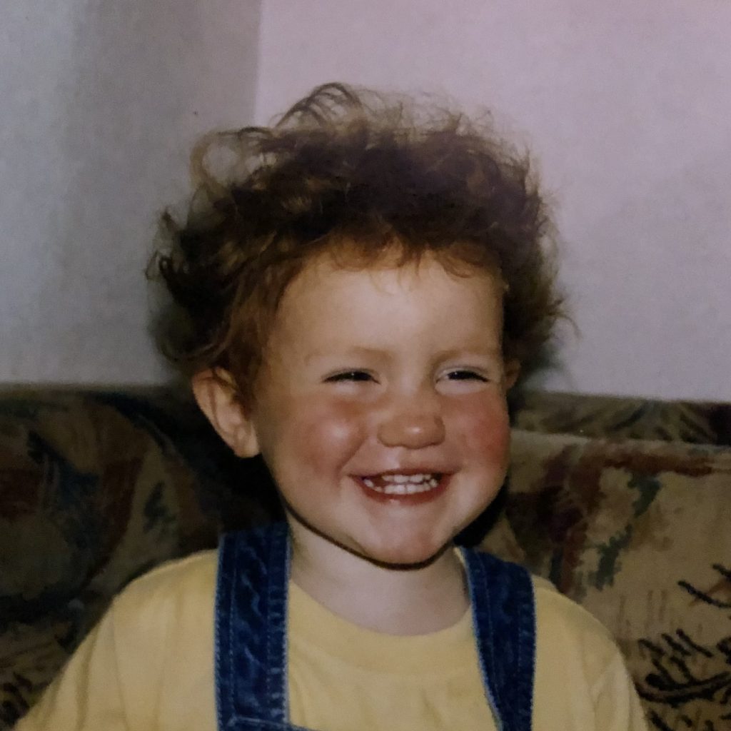 Heather Cartwright (Janey) as a child with short curly hair wearing a yellow top and blue dungarees sat on a patterned sofa.