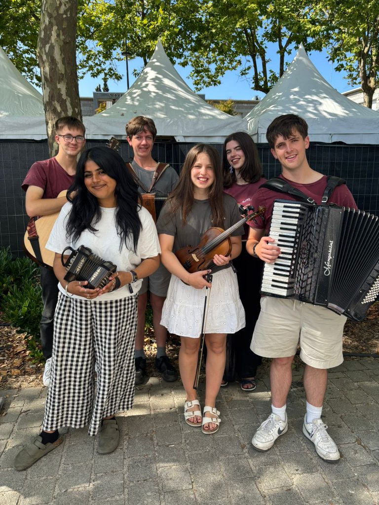 Fèis Rois Cèilidh Trail young traditional musicians posing outdoors with their instruments, including guitar, fiddle, accordion, and concertina, at a summer music festival.