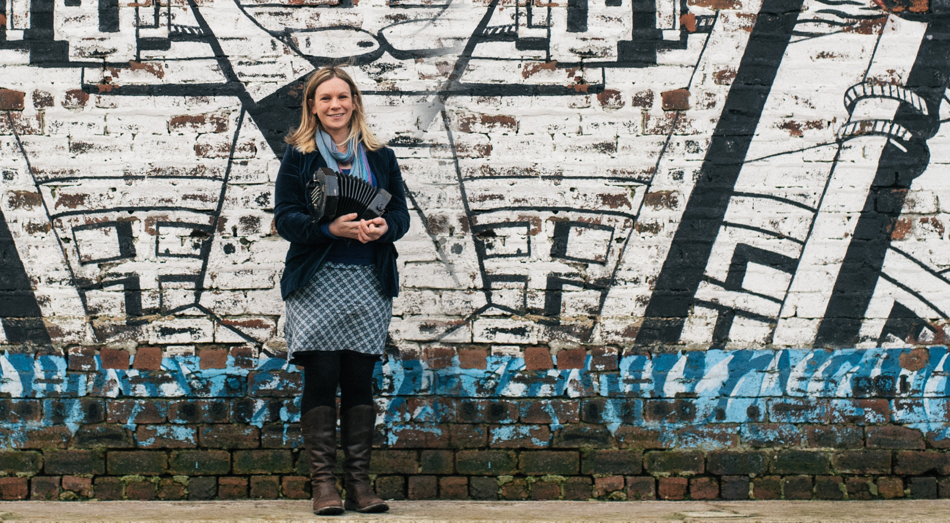 Frances Wilkins stands in front of a painted wall, holding a concertina in her arms. With blonde hair she is wearing a black jacket and a blue checked skirt and smiling.