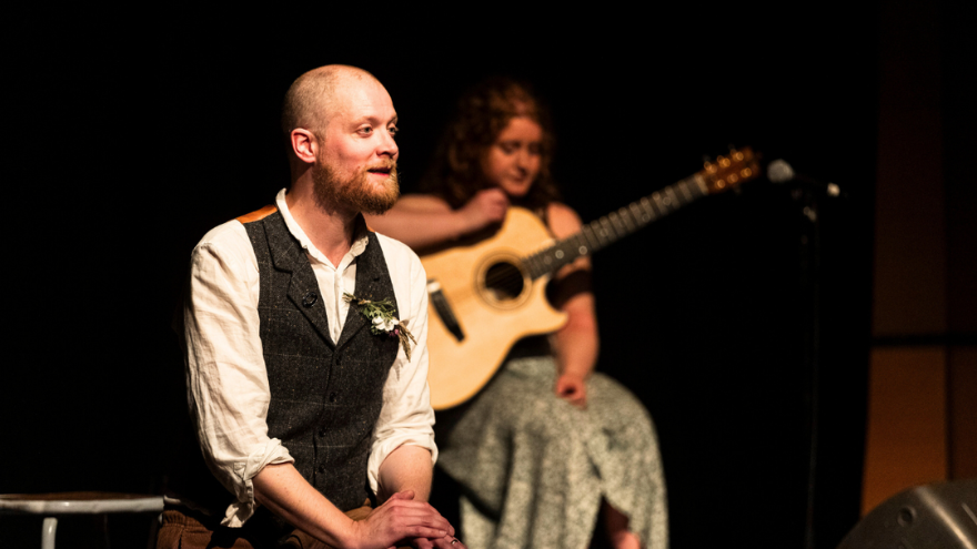 storyteller Daniel Serridge with musician in the background, sitting down telling a story