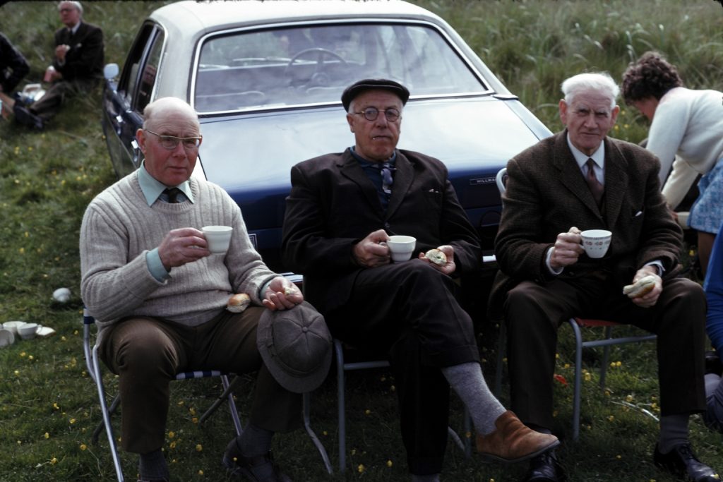 Three old men sit in front of a car on chairs on the grass, drinking tea and eating sandwiches.