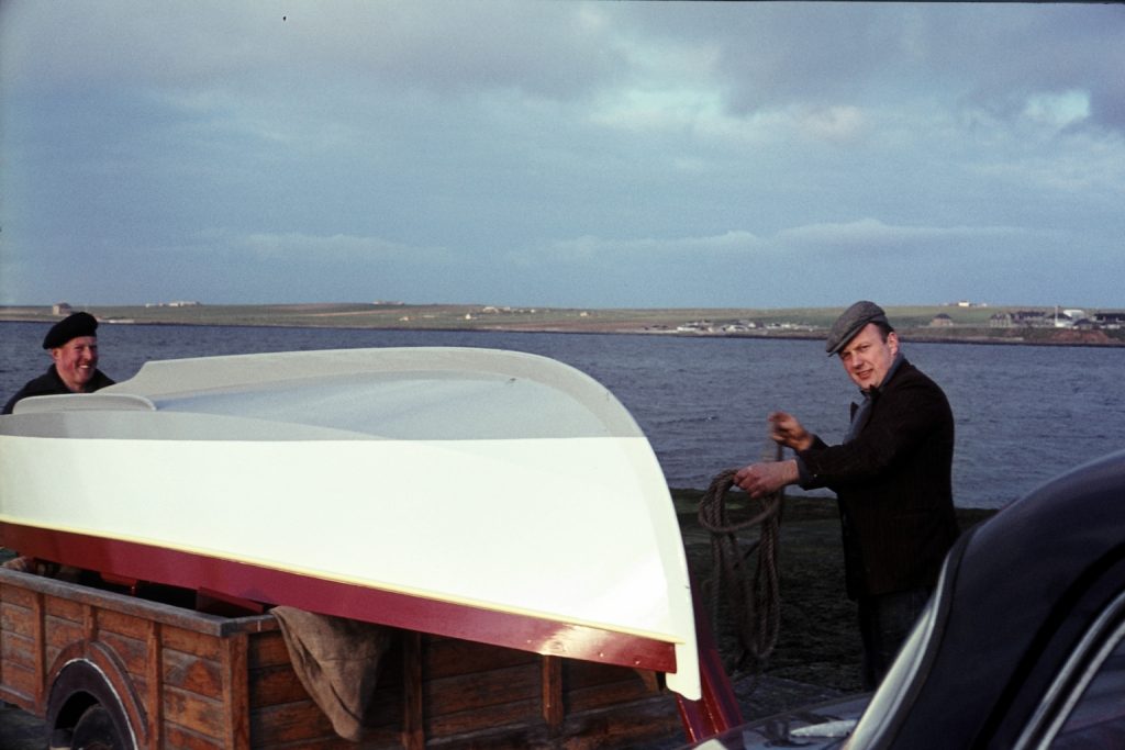 Two men on a small boat smiling at the camera, with the sea and land in the background.