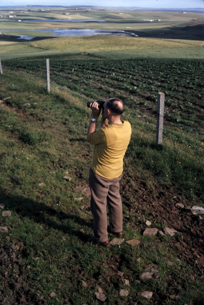 A man in a yellow t shirt stands in a field holding binoculars up to his eyes.