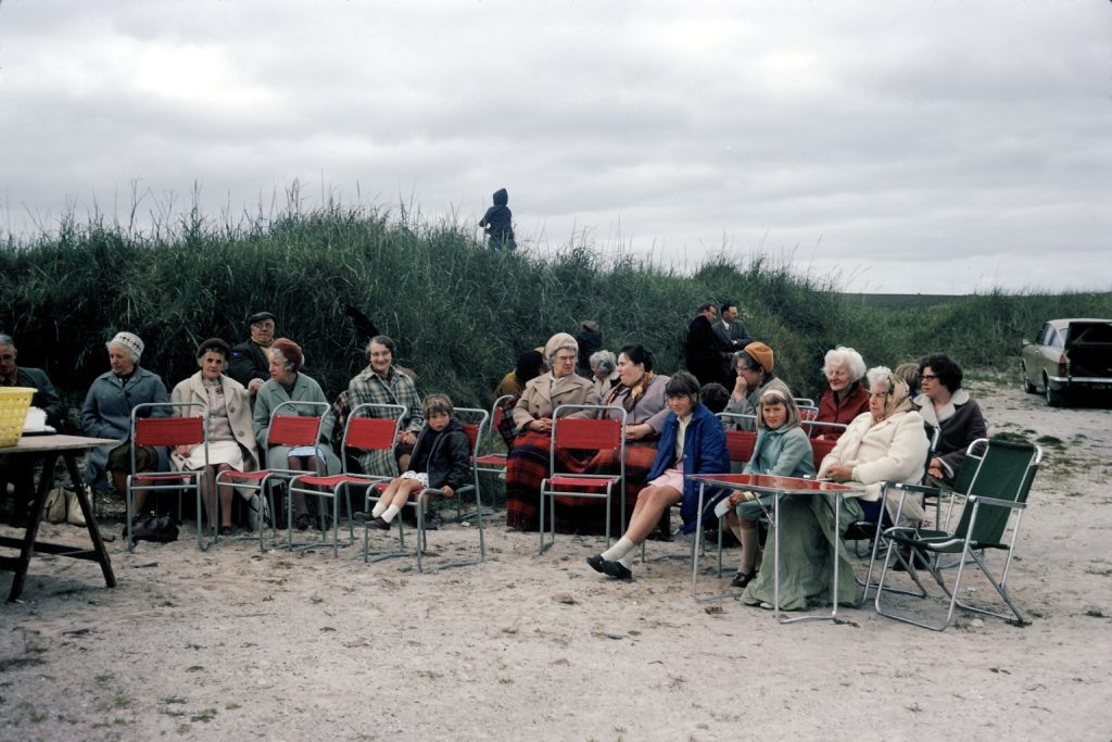 A group of mainly women and children sit on red chairs on the beach, with long grass and a car in the background.