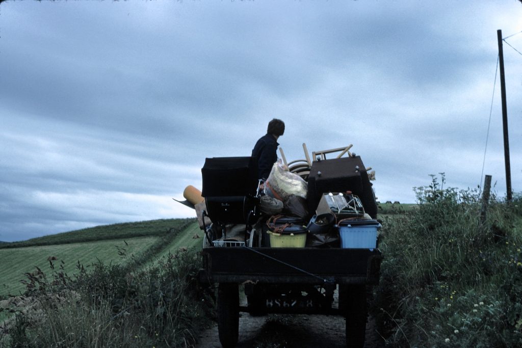 A man on a tractor filled with brick-a-brack on a country lane with hedges and fields.