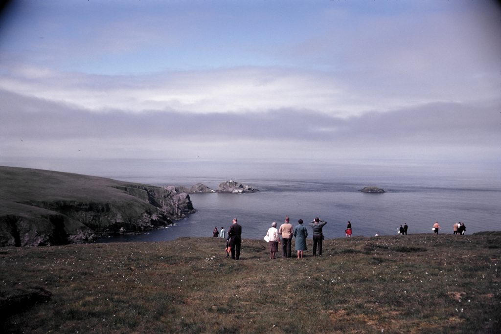 a group of people stand on the clifftop overlooking the sea, on a sunny and cloudy day.