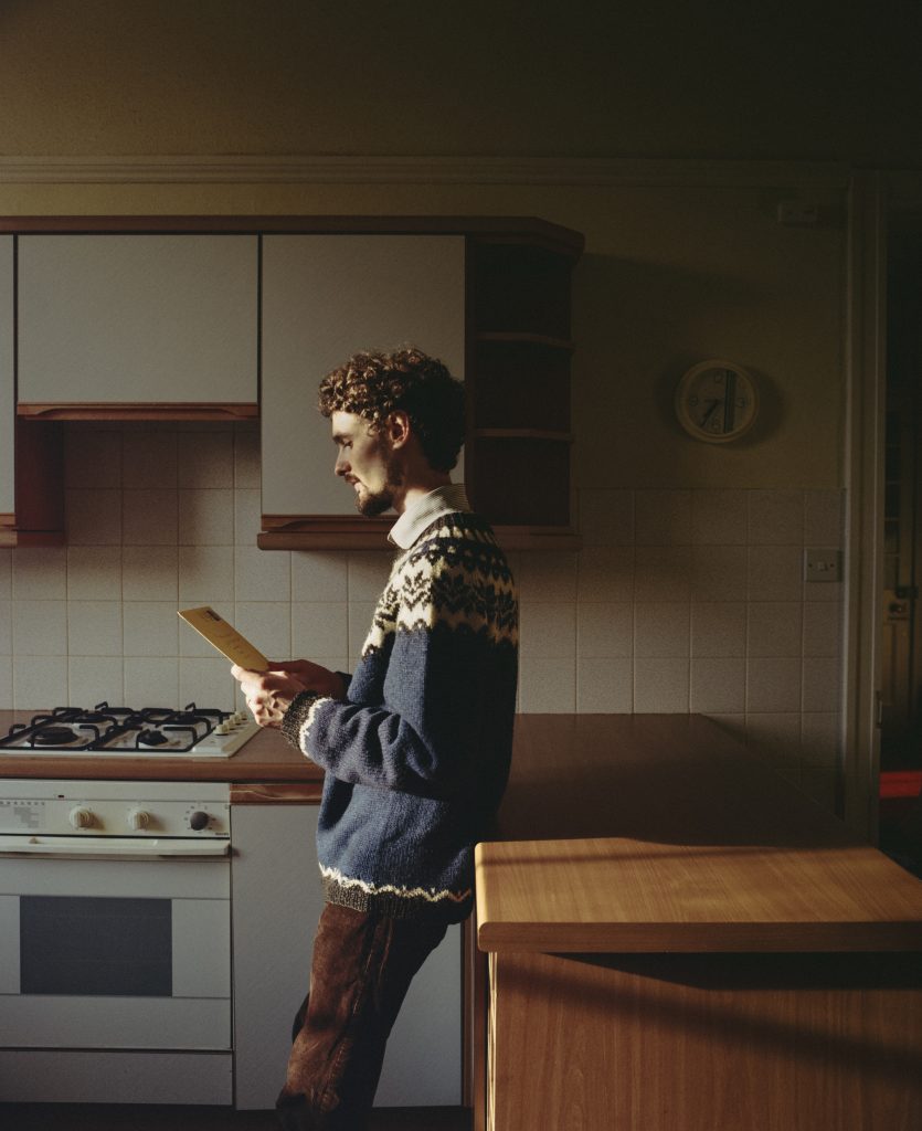 Chris Amer stands in a kitchen wearing a knitted jumper and looking pensively at an envelope.