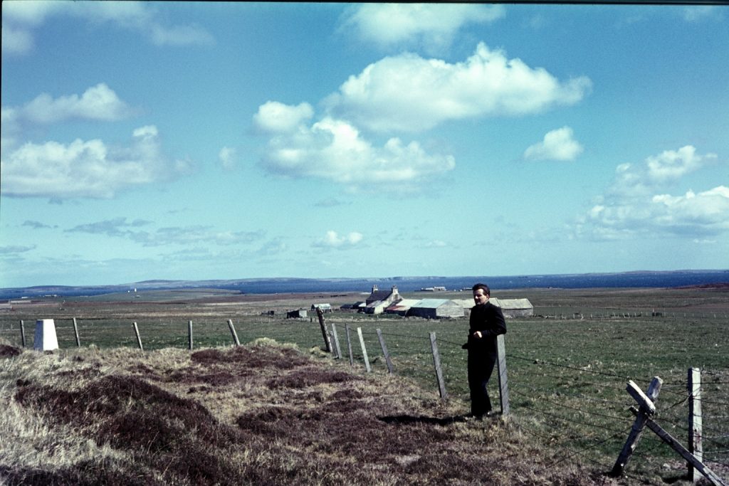 A man stands in front of a fence with a field and farmhouse in the background, on a sunny and cloudy day.
