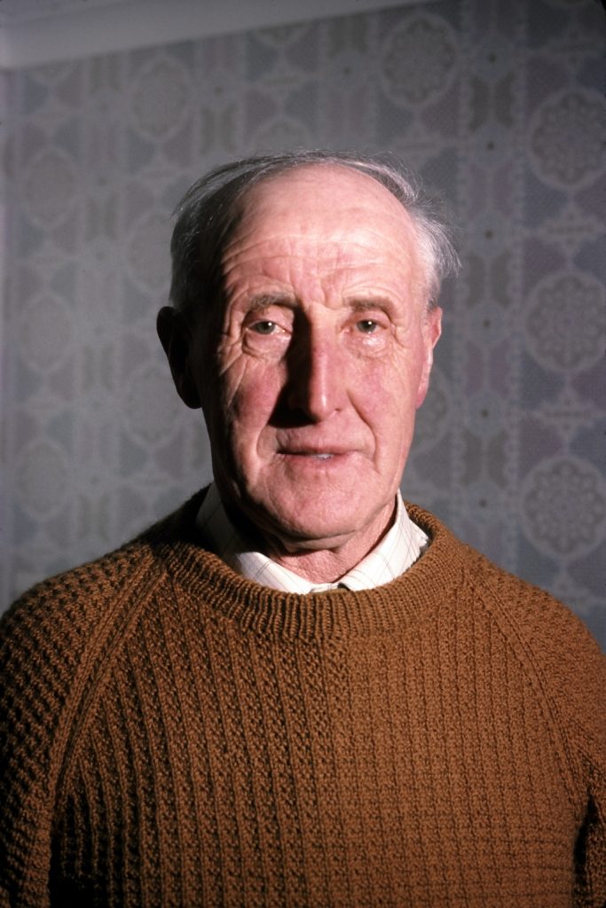 A portrait of an old man smiling at the camera wearing a knitted burn orange jumper with patterned wallpaper in the background.