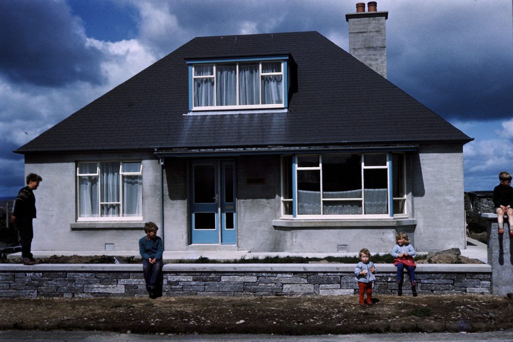 Five children stand and sit on a wall outside a house in the sunlight.
