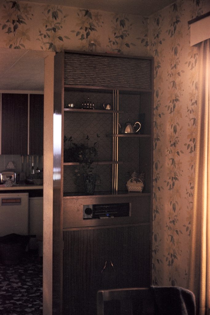 An old photo of a kitchen dresser with a silver teapot, with leafy wallpaper in a 1970s style.
