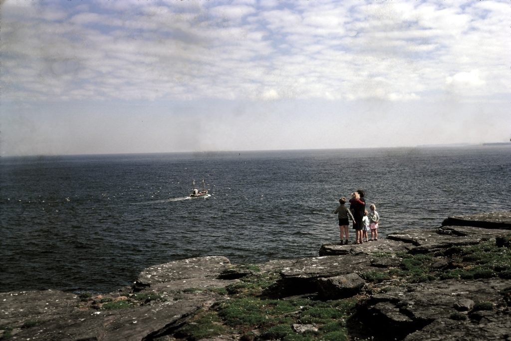 A grandmother and her four grandchildren stand on rocks looking out to sea and the horizon. There is a boat in the sea and clouds in the sky, but the sun is shining.