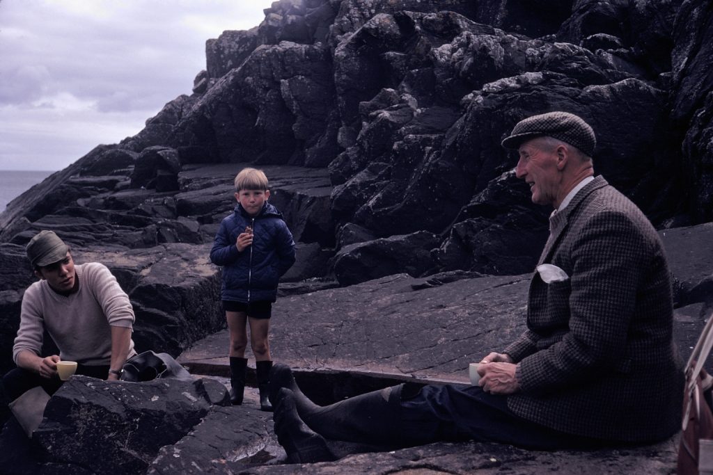 Two men and a child sit and stand on rocks drinking tea and eating snacks.
