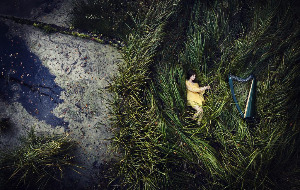 A drone photo taken from above of Lucie Hendy lying down in a yellow dress in the long green grass, with her harp laid down next to her.