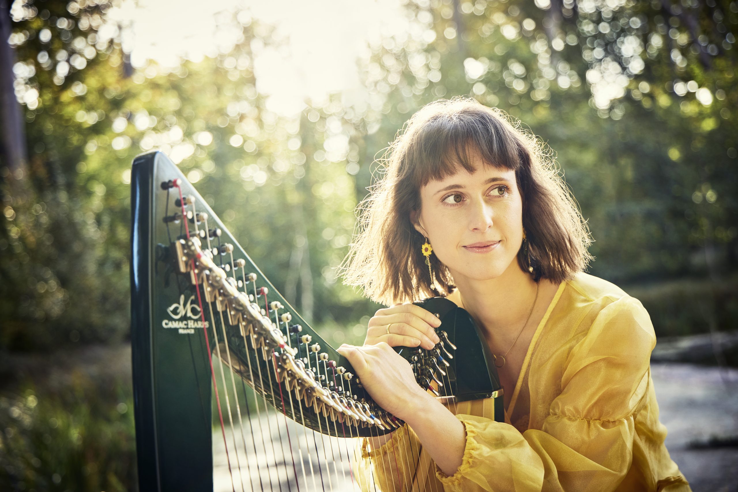 Lucie Hendry sits in a yellow dress leaning on her harp, with a background of trees.